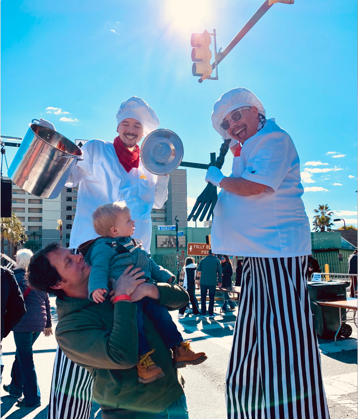 Two chefs on stilts entertain a child held by a man outdoors. The chefs wear white coats, chef hats, and striped pants.