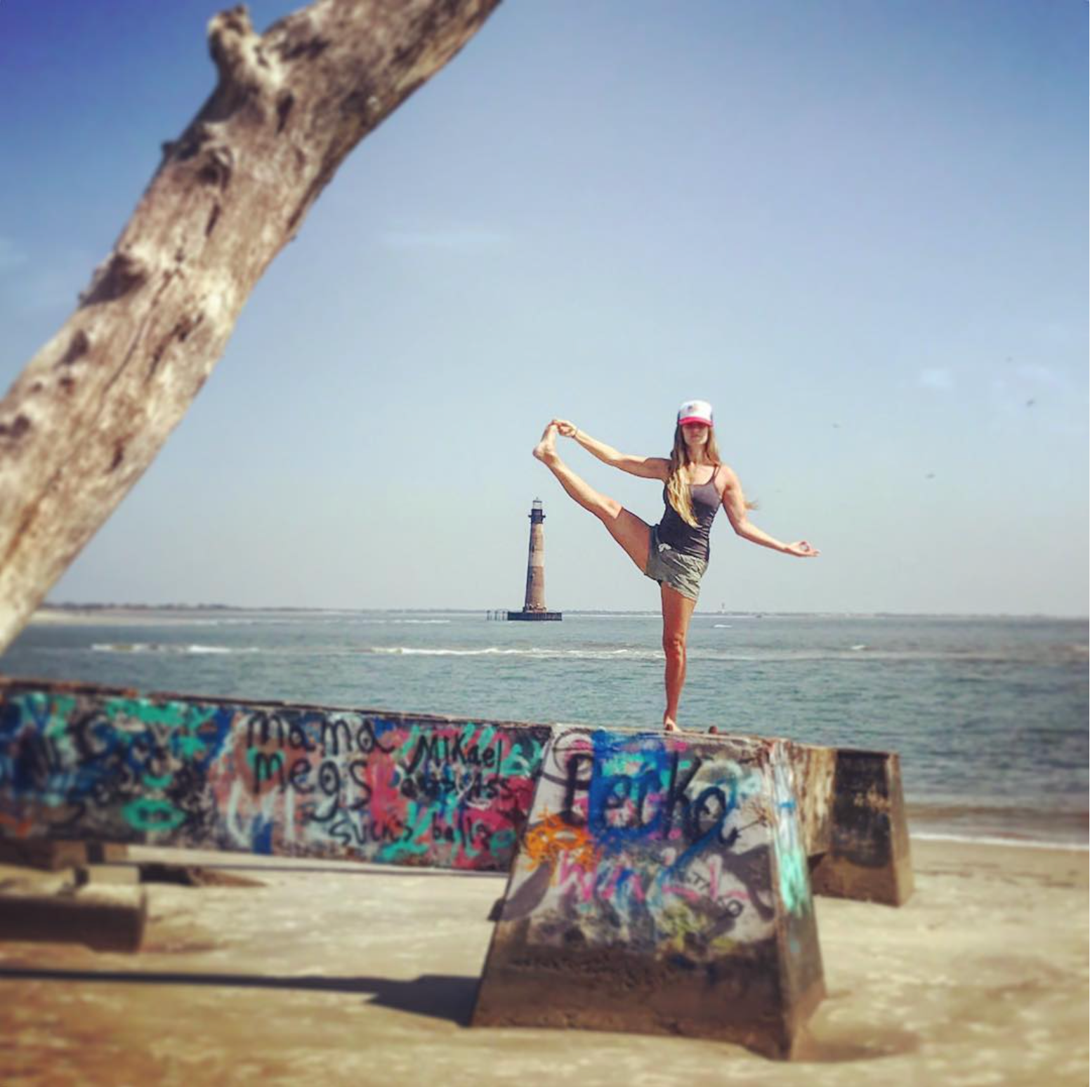 Woman in yoga pose on a graffiti-covered wall at the beach, with a lighthouse in the distance.