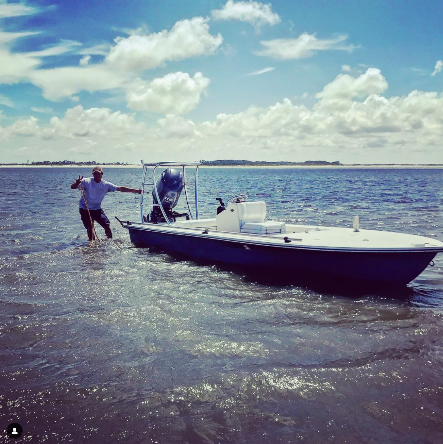 Man pulling a blue boat through shallow water towards a distant island on a sunny day.