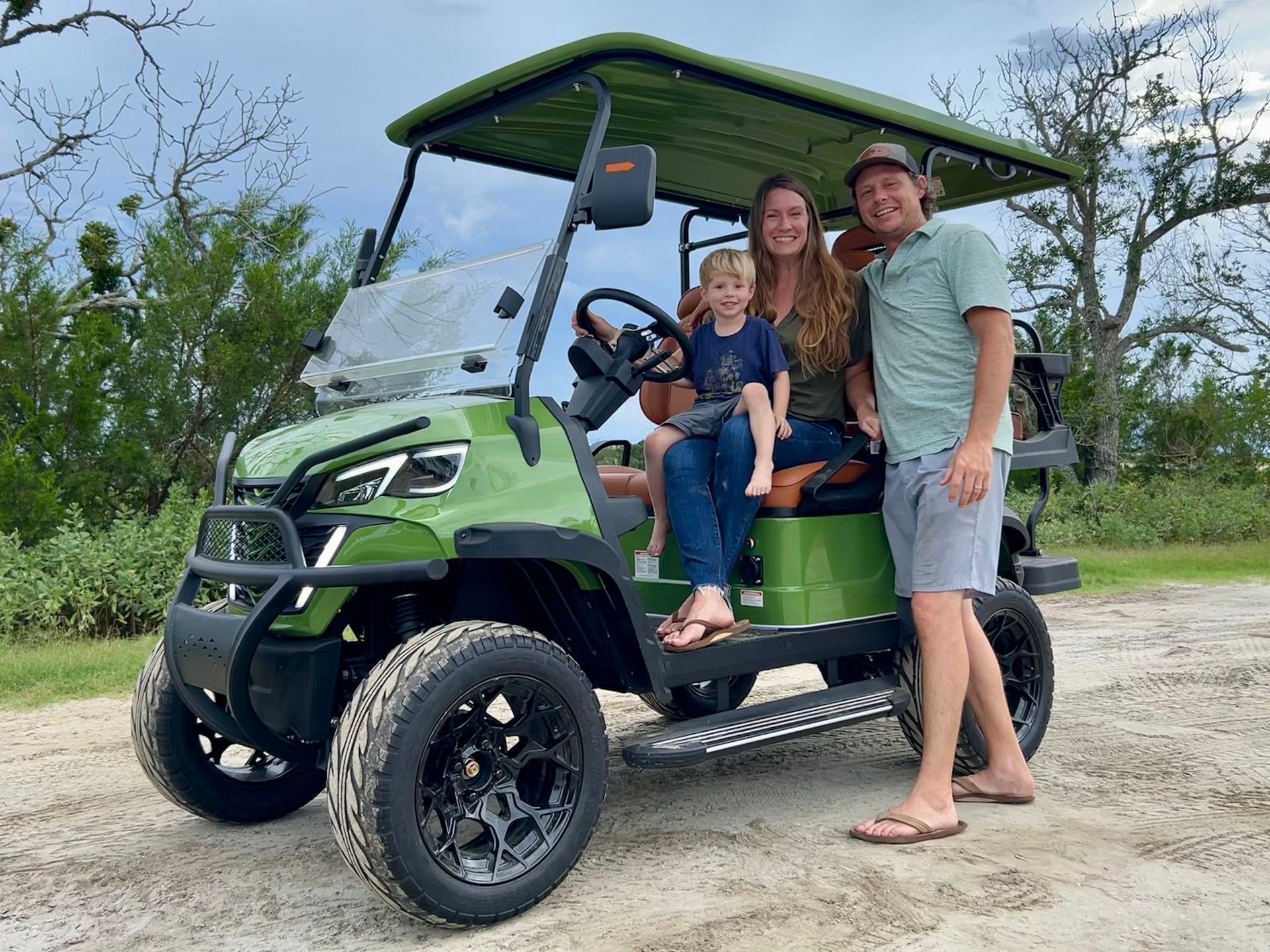 Family poses with a green golf cart on a gravel road; they smile in front of a natural backdrop.