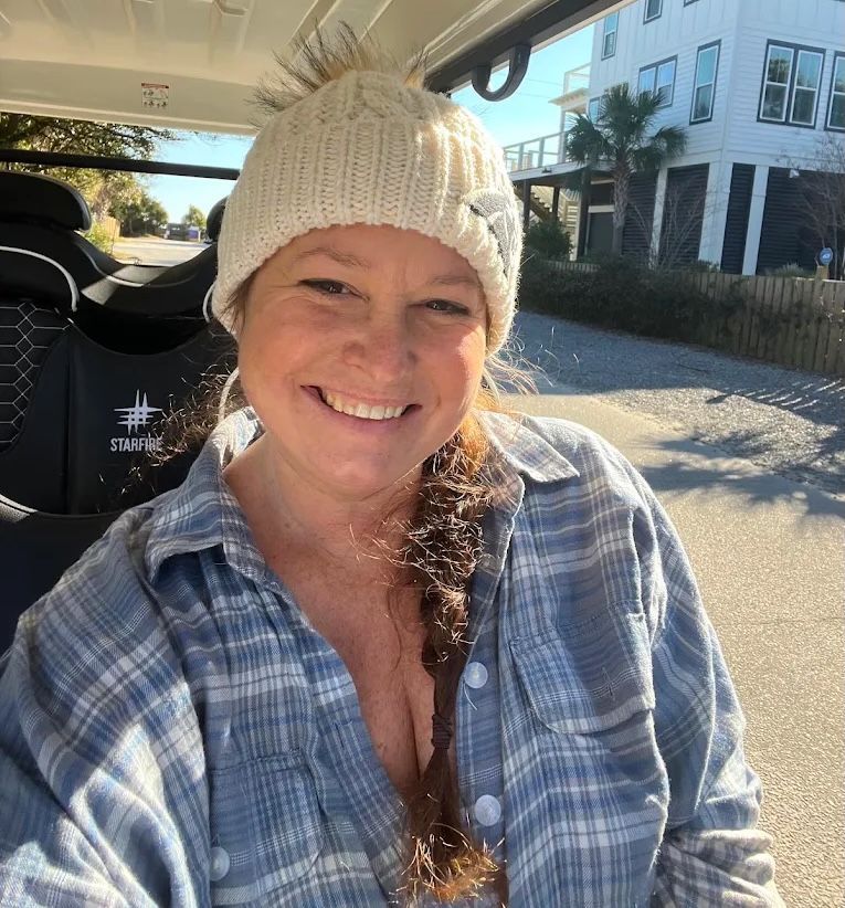 Woman in a golf cart smiling, wearing a white hat and blue plaid shirt. She has a long braid and is outdoors on a sunny day.