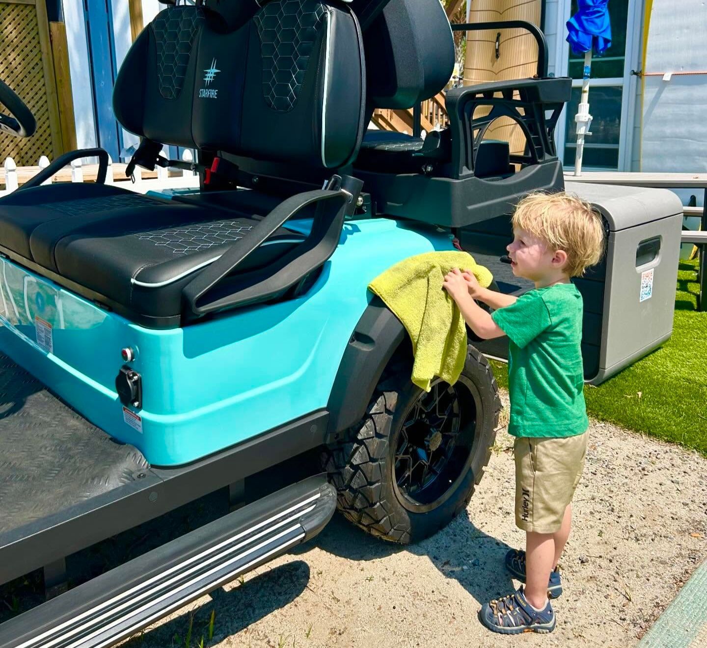 Young child in a green shirt cleans a light blue golf cart tire with a yellow cloth outdoors.