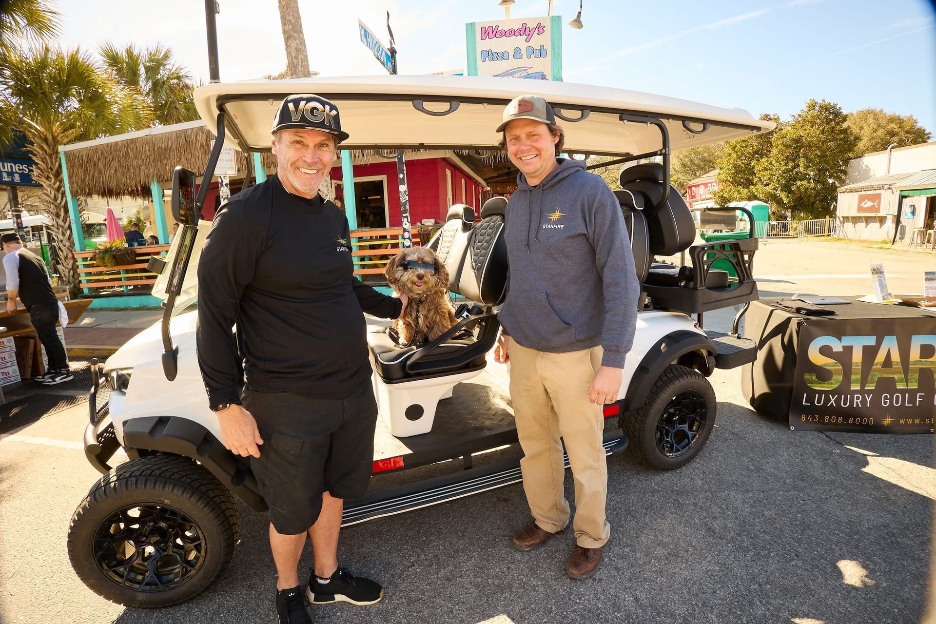 Two men and a dog pose beside a white golf cart with black wheels in front of a shop. The dog sits in the driver's seat.