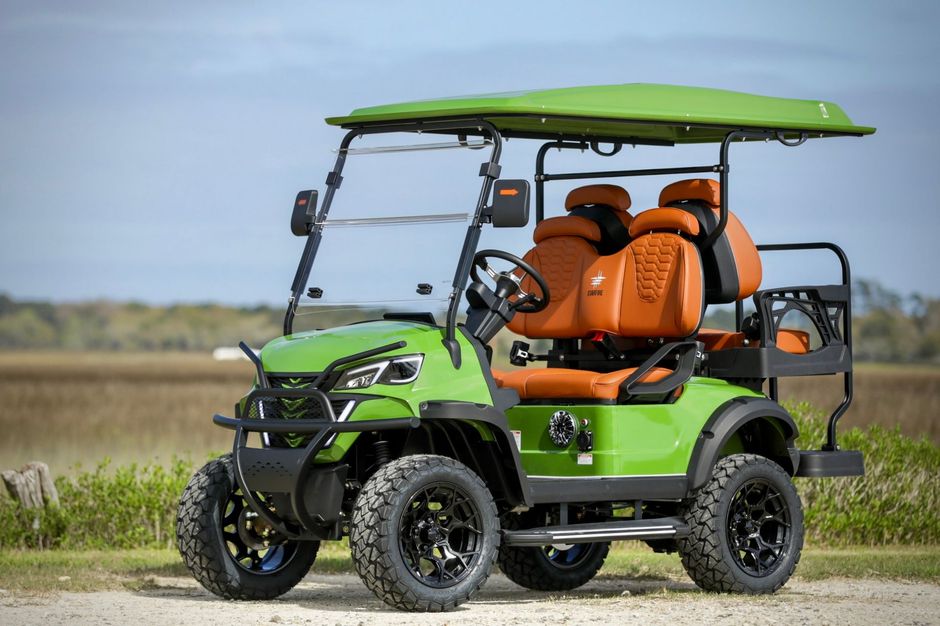 Green golf cart with orange seats and black wheels in a field on a sunny day.