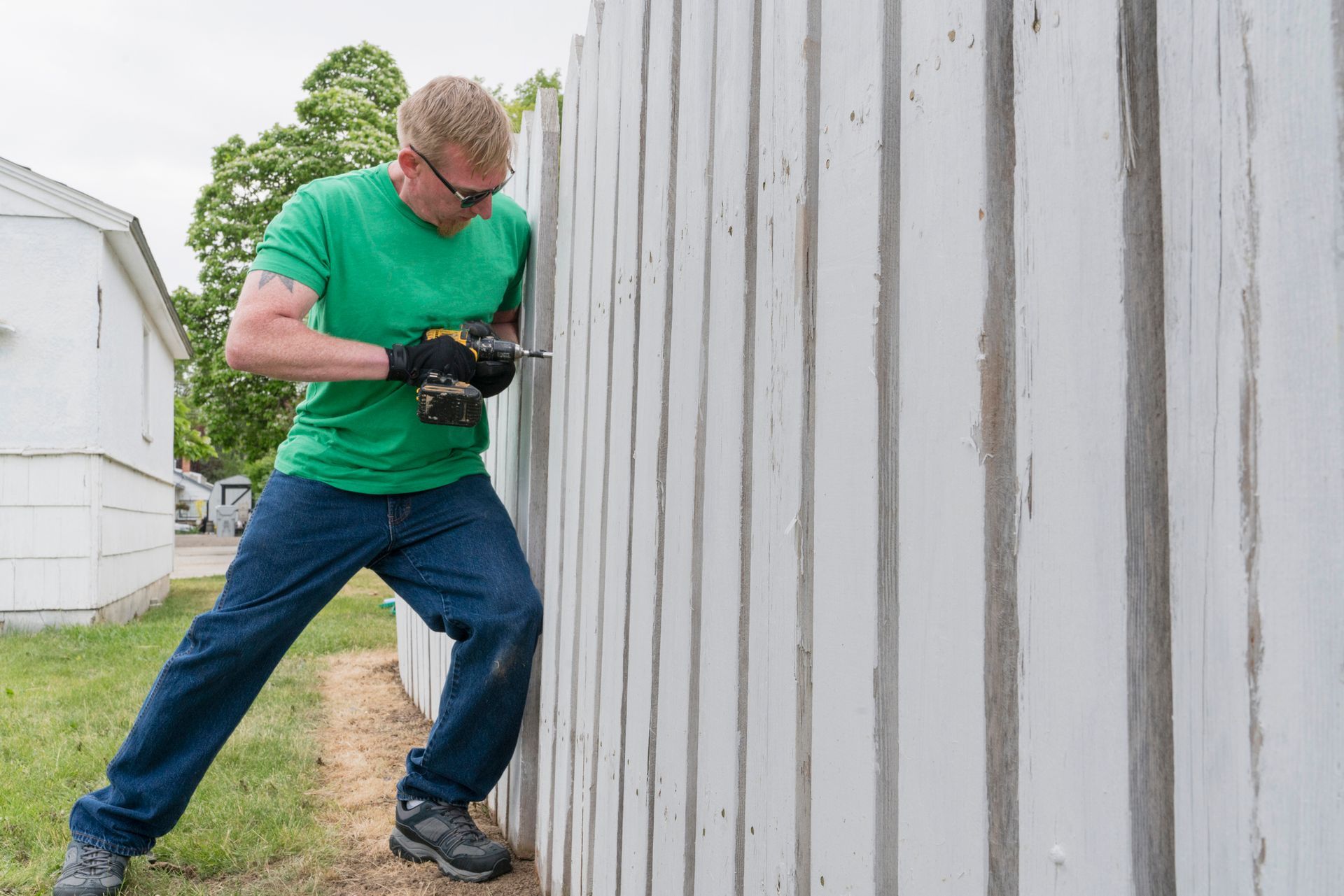 Man in green shirt and jeans using a drill on a white wooden fence outdoors.