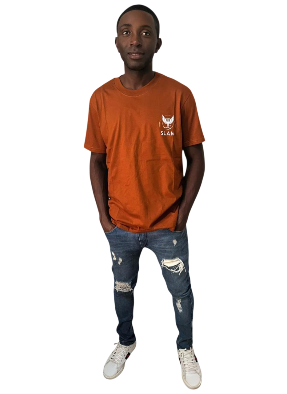 Hombre con una camiseta naranja y jeans rotos, con las manos en los bolsillos, de pie y con una expresión neutra.