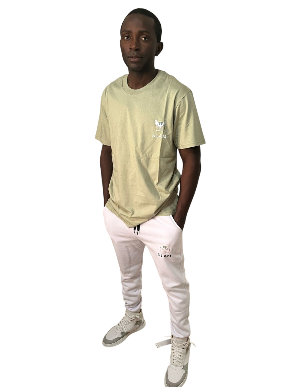 Hombre con camiseta color crema y pantalón negro, con las manos en los bolsillos, de pie y sonriendo.