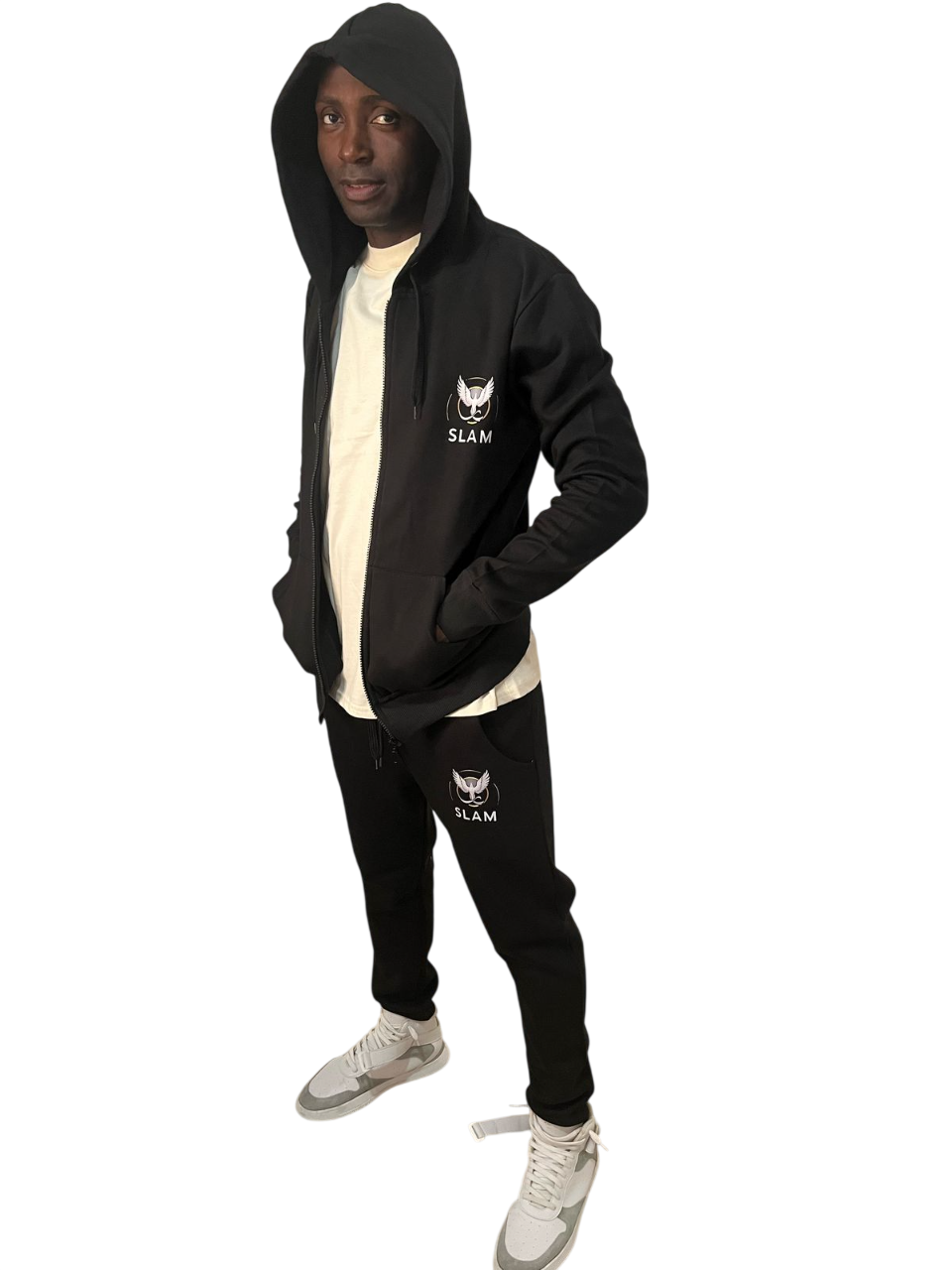 Persona de pie con chándal negro con capucha. Logotipo blanco en el pecho, pantalones y zapatillas claras.