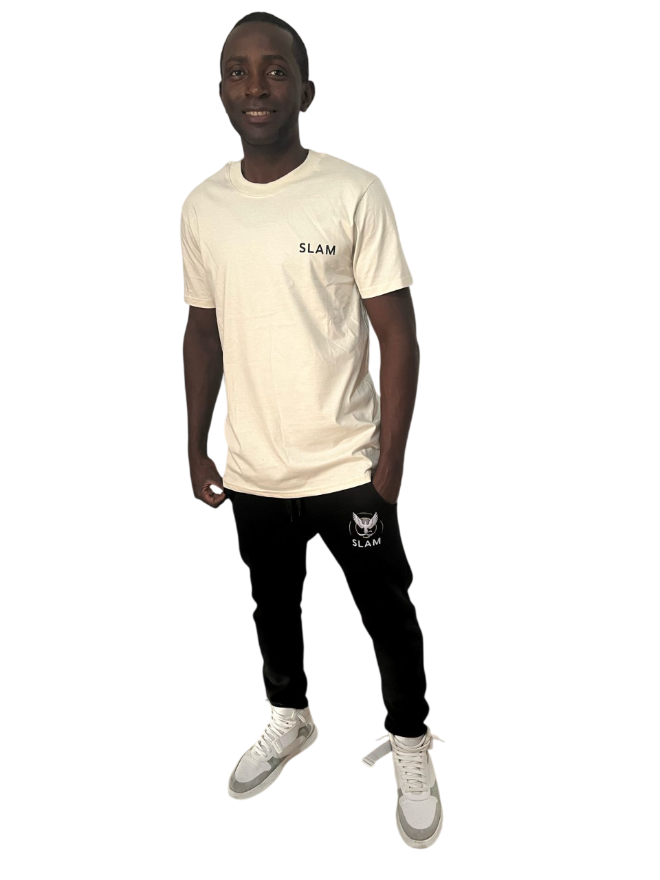 Hombre con camiseta color crema y pantalón negro, con las manos en los bolsillos, de pie y sonriendo.