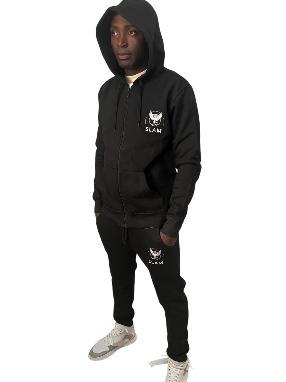 Persona de pie con chándal negro con capucha. Logotipo blanco en el pecho, pantalones y zapatillas claras.