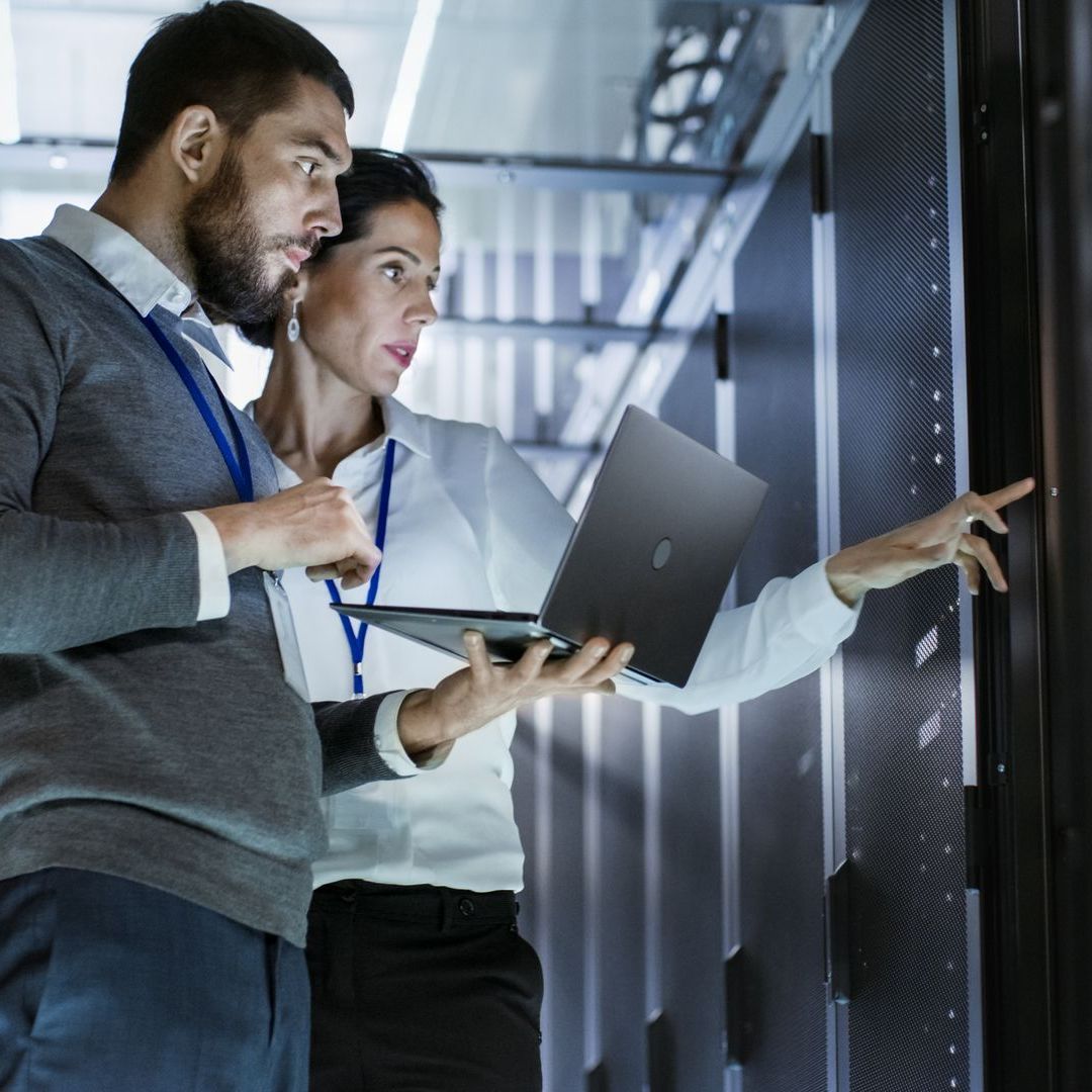 A man and a woman are looking at a laptop in a server room.