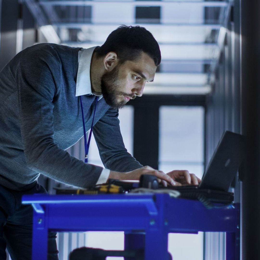 A man is working on a laptop computer in a server room.