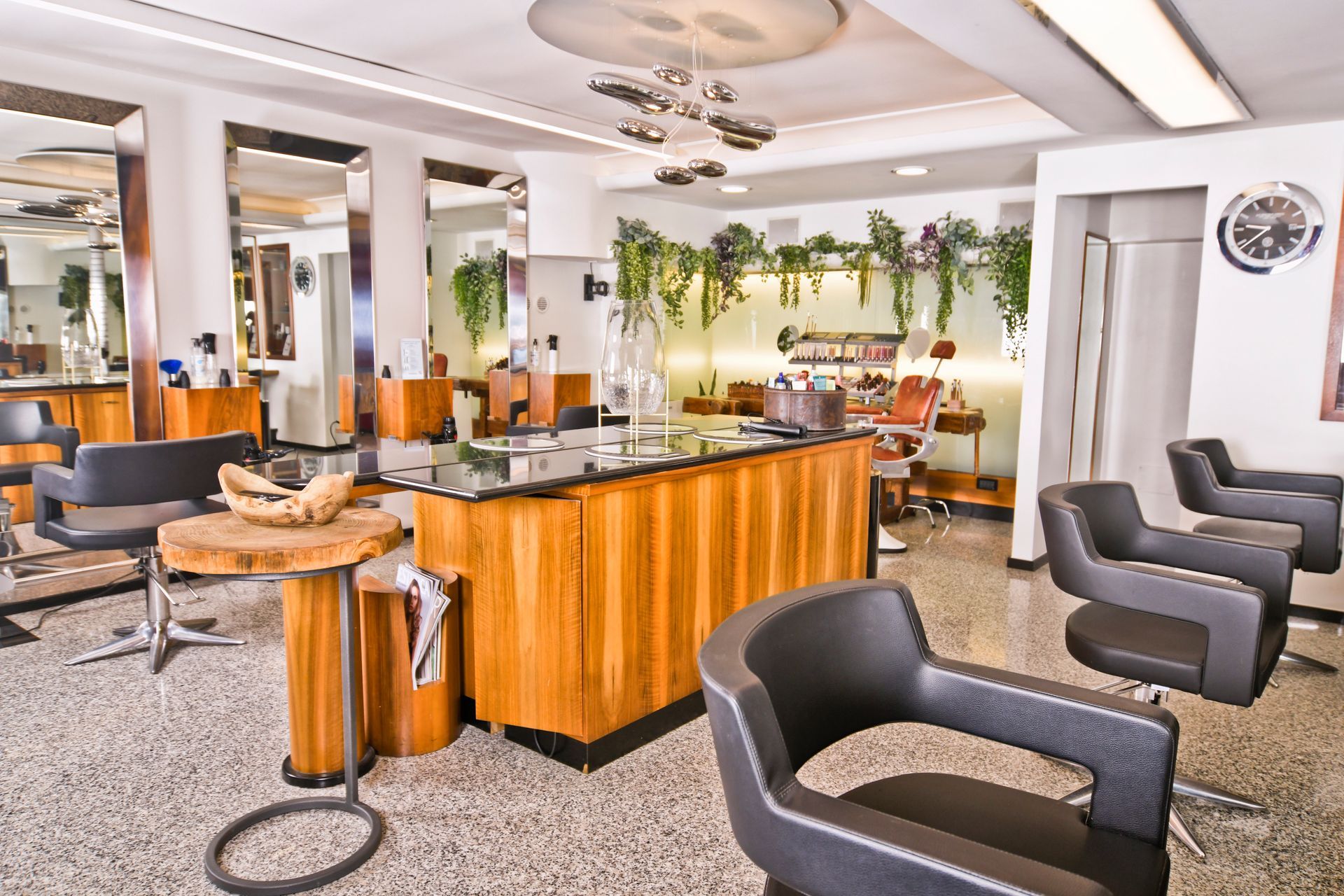 Interno di un salone da parrucchiere con postazioni per lo styling, specchi, mobili in legno e piante sospese.