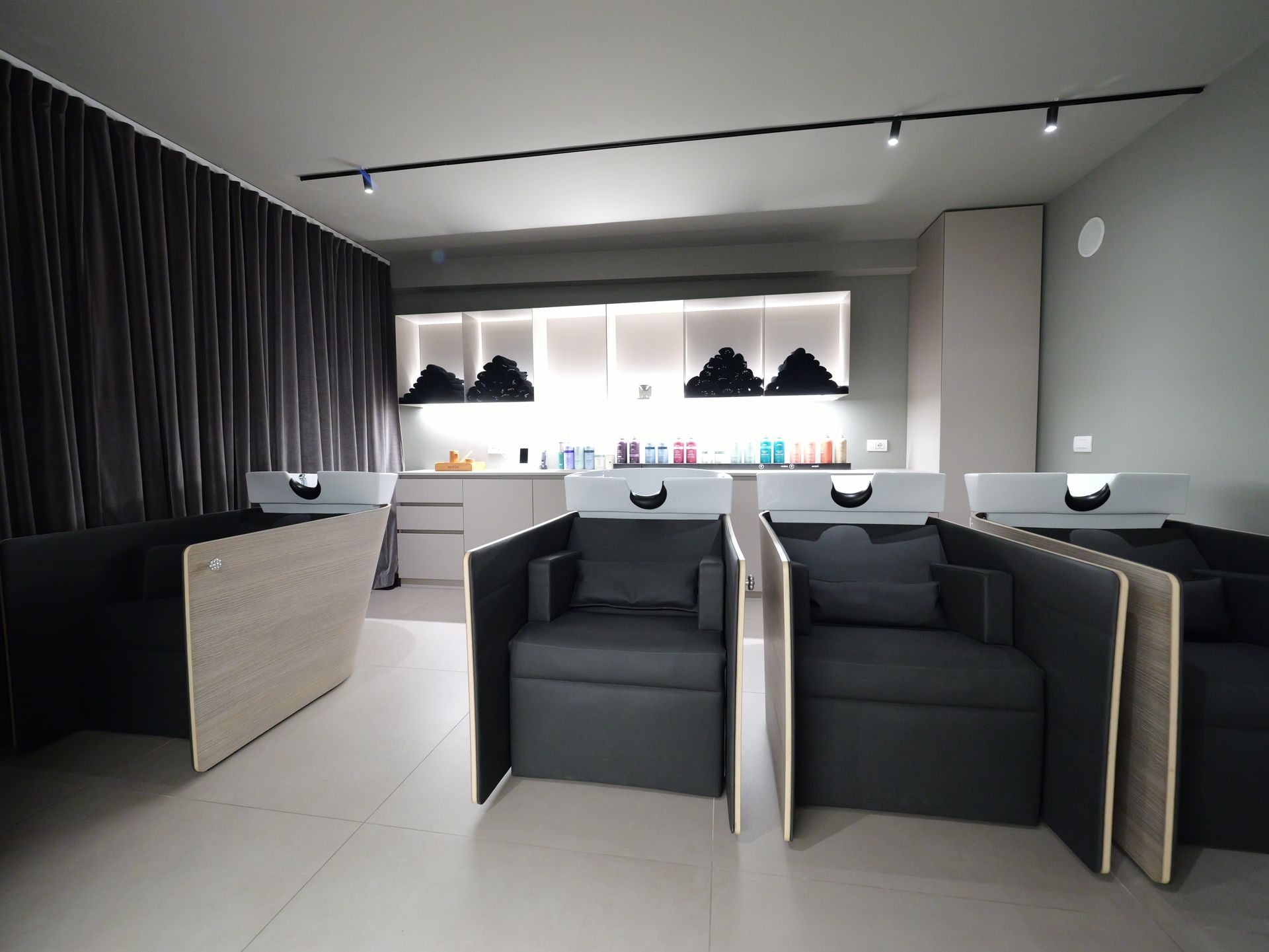 Postazioni di lavaggio del salone con poltrone e lavandini scuri, pareti grigie e luci intense dall'alto.