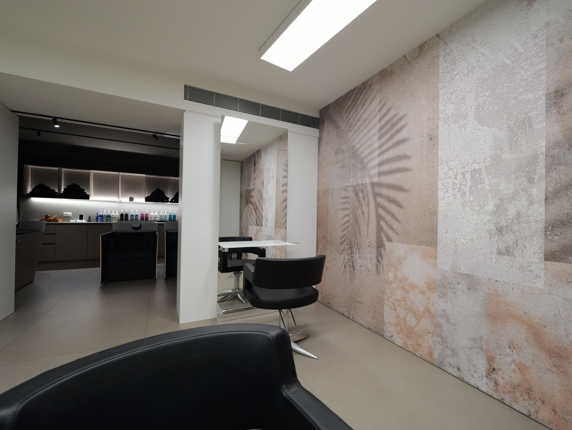Interno di un salone da parrucchiere con poltrone, specchio e parete strutturata con motivo a foglie.