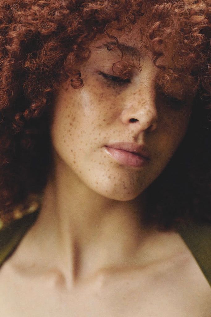 Donna con capelli ricci castano-rossicci, pelle lentigginosa, occhi chiusi, sguardo rivolto verso il basso.