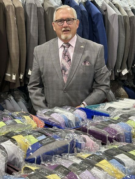 Man in a grey suit stands behind a table of shirts. Suits hang in the background.