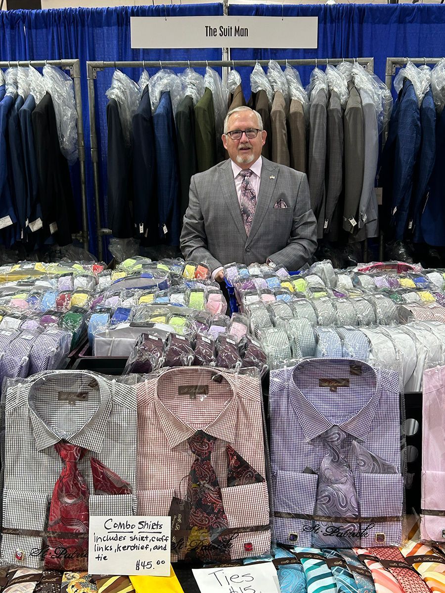 Man in suit stands at clothing booth, shirts and suits for sale.