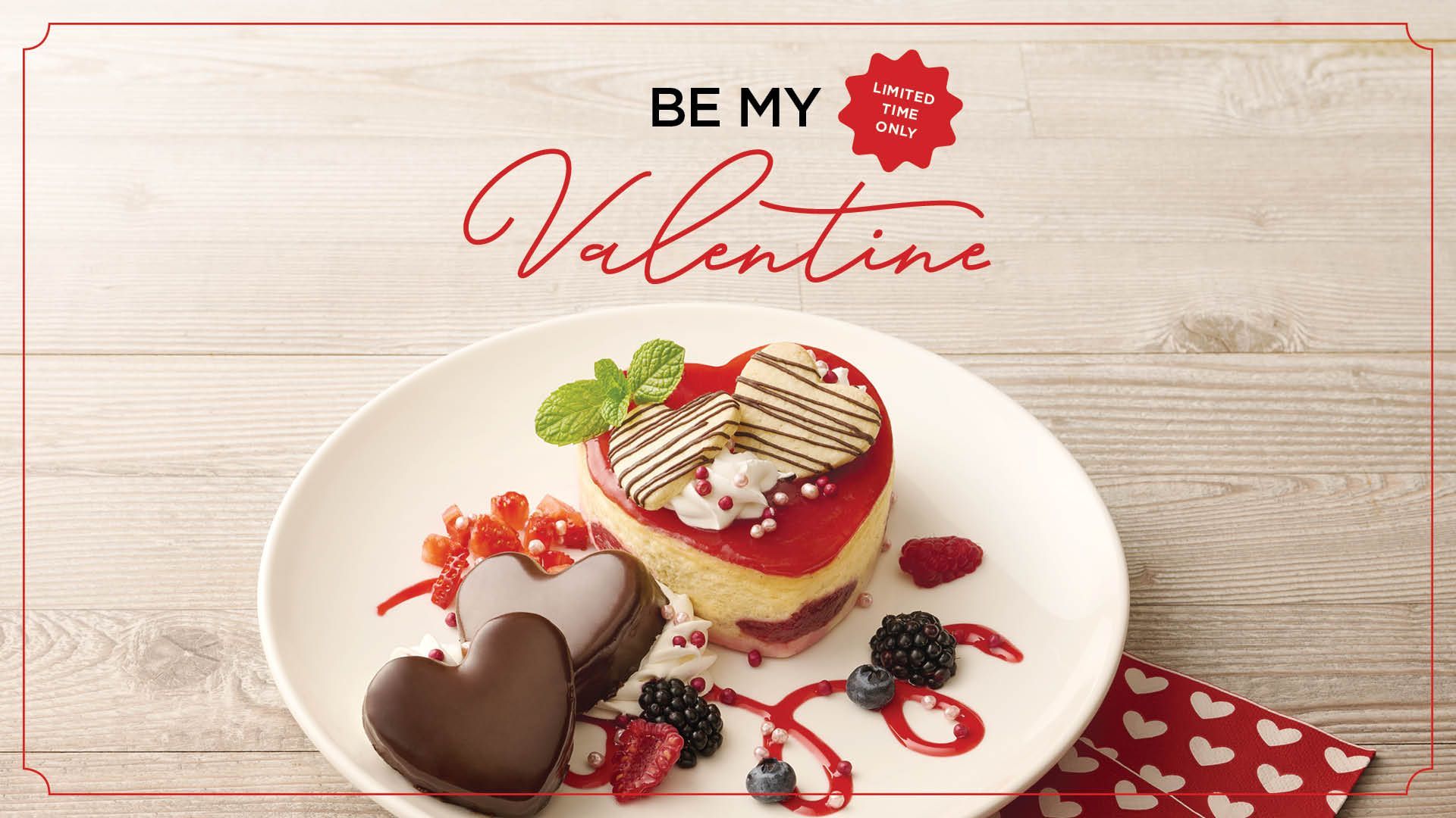Valentine's Day dessert: heart-shaped cake, chocolate hearts, berries, and sauce on a white plate with a festive napkin.