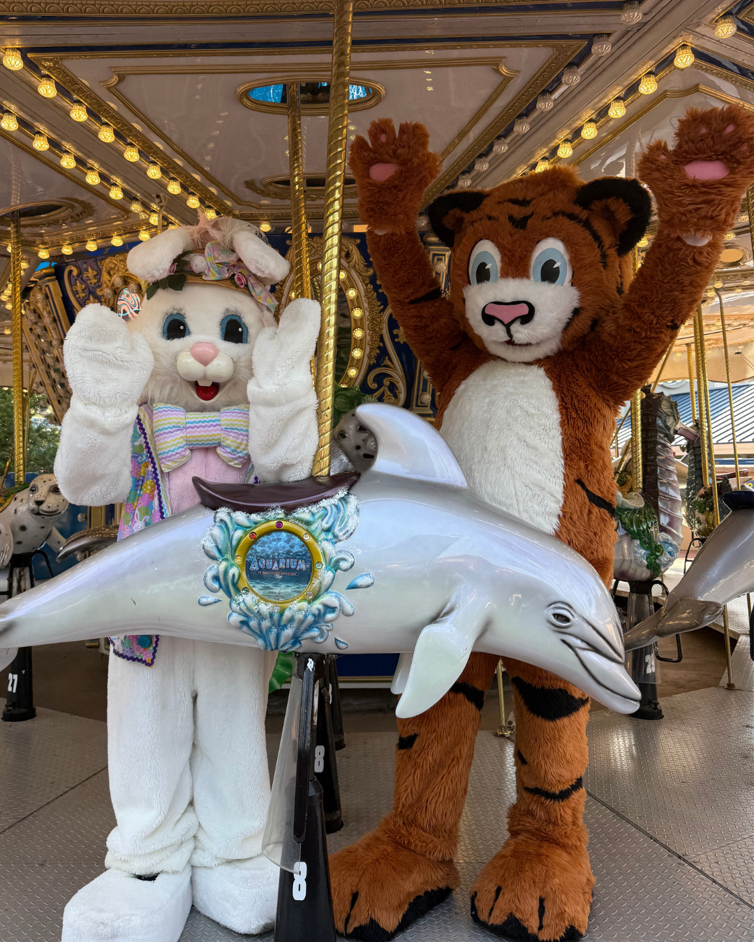 A bunny and tiger mascot standing on a carousel, each with arms raised, by a silver carousel dolphin.