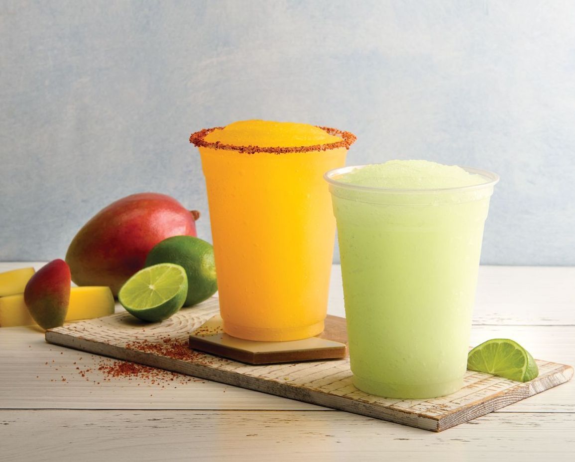 Two drinks on a cutting board with fruit in the background