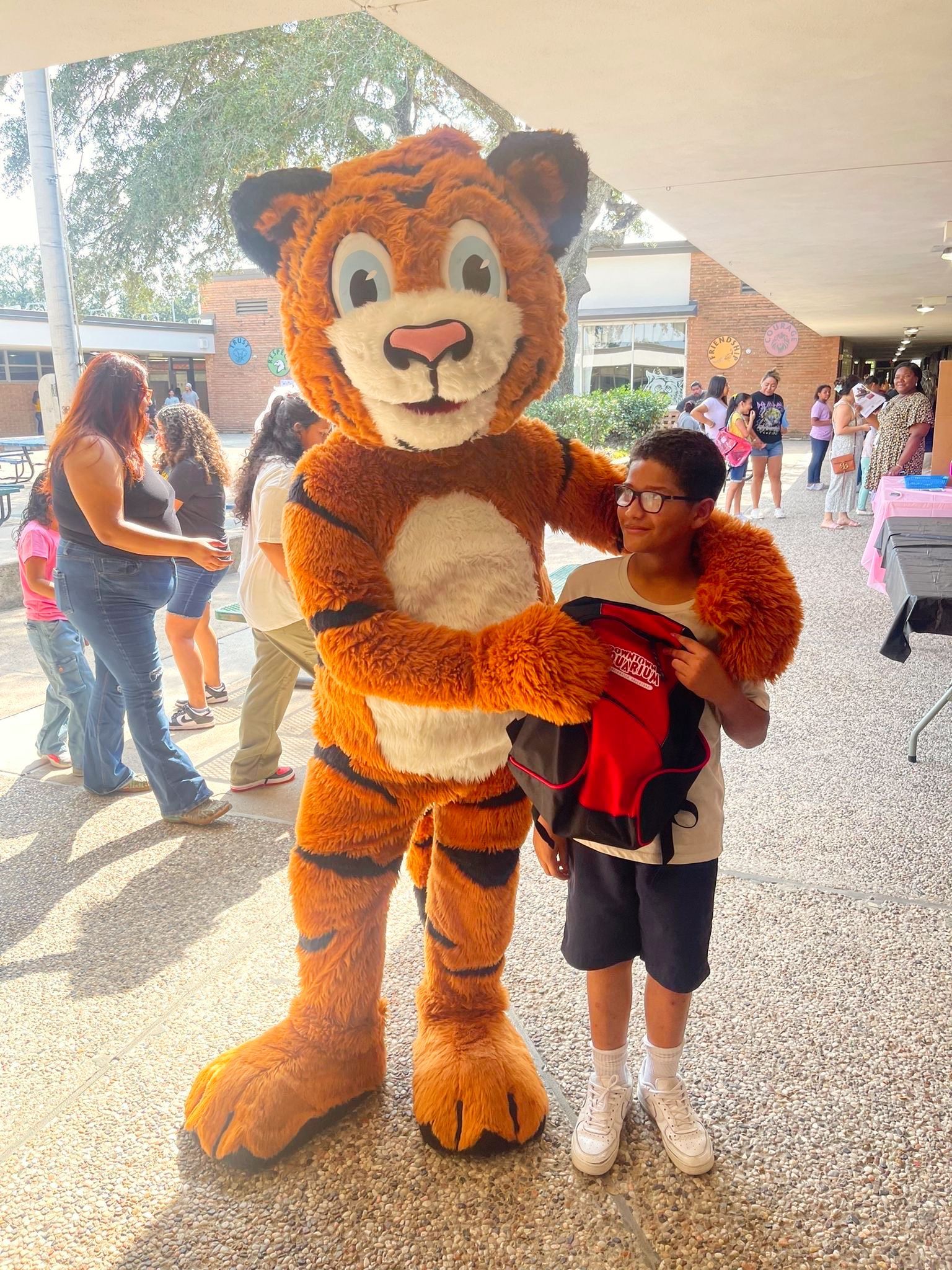 A boy holding a red backpack stands next to a tiger mascot