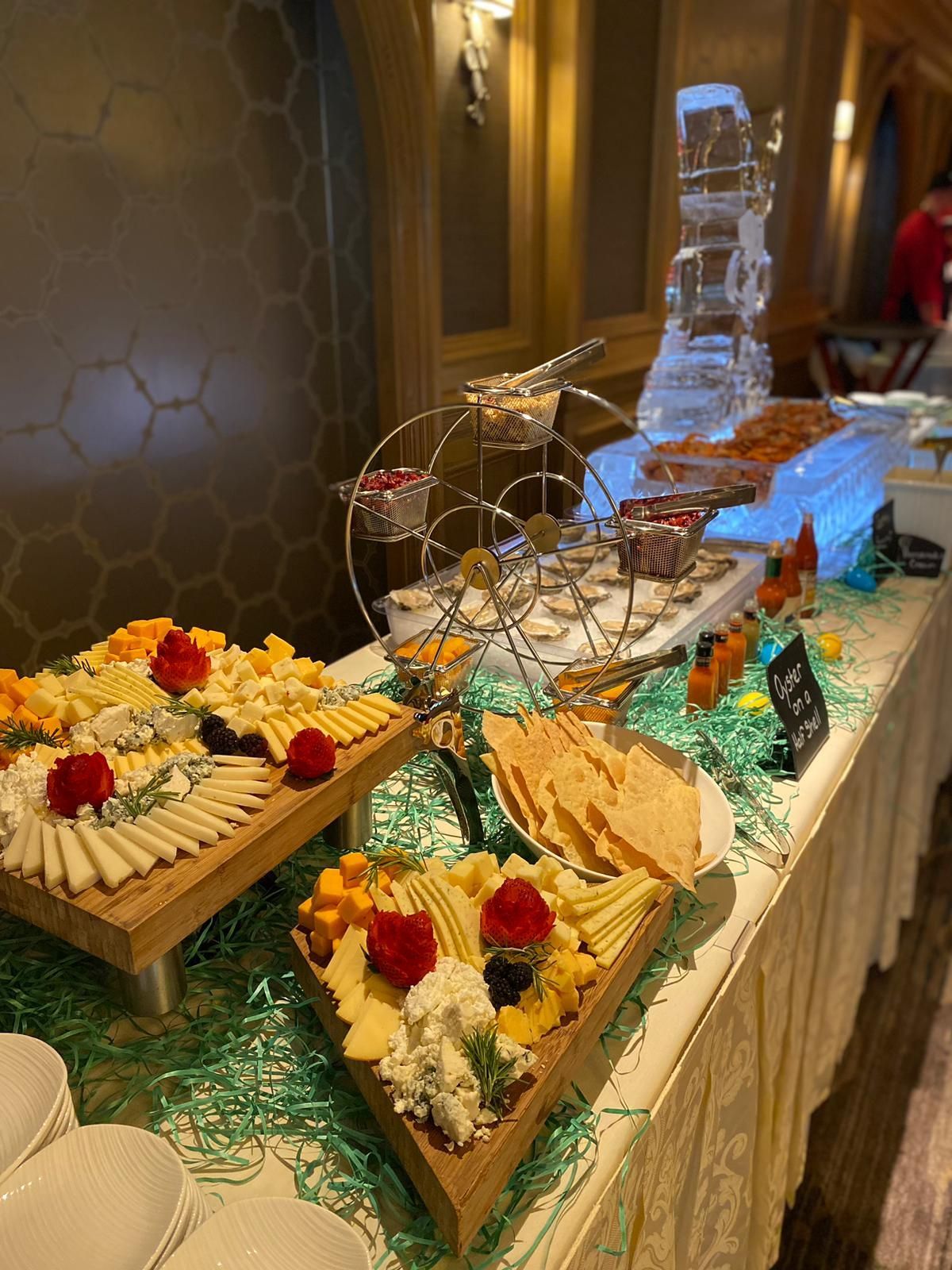 A buffet table features wooden platters of assorted cheeses, crackers, a rotating display stand, and an ice sculpture.