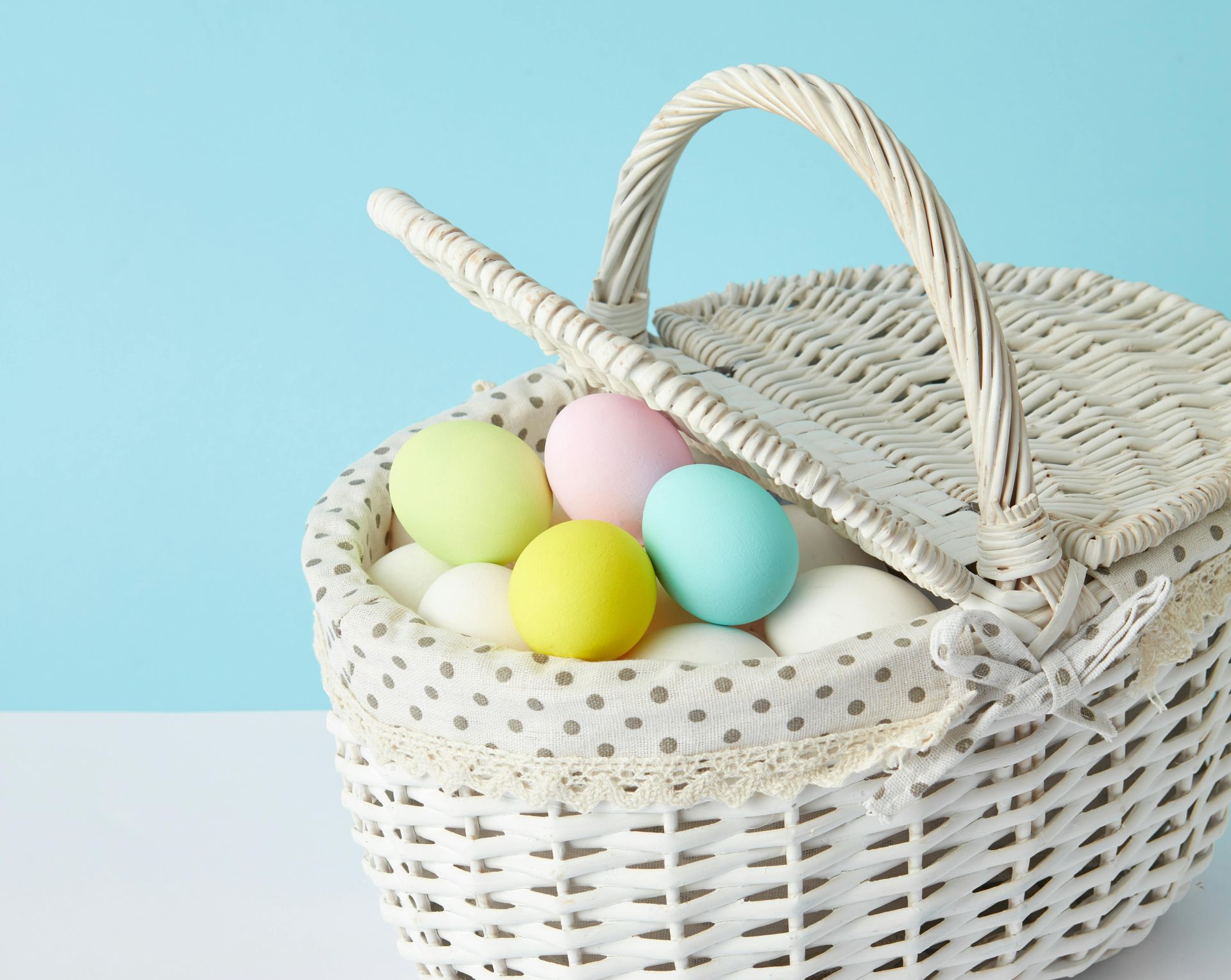 A white wicker basket filled with colorful pastel Easter eggs, set against a light blue background.