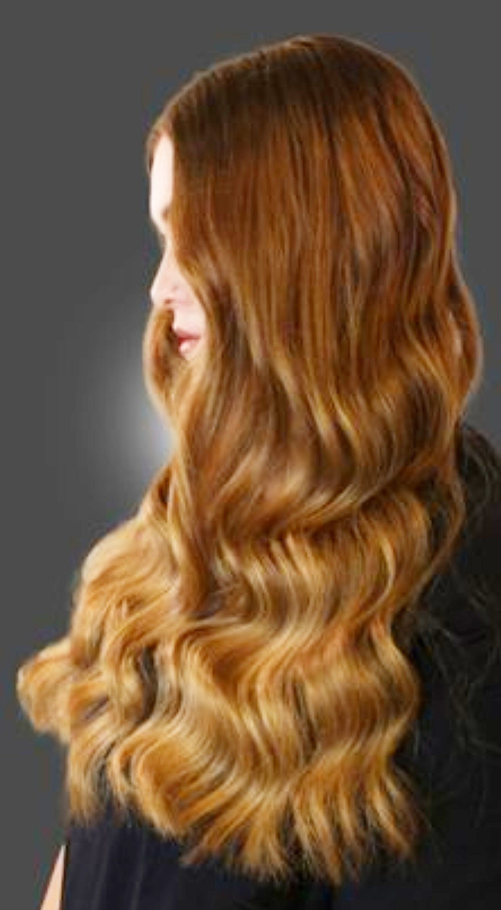 Una vista di profilo di lunghi capelli ondulati color caramello che ricadono lungo la schiena di una persona, su uno sfondo grigio scuro.