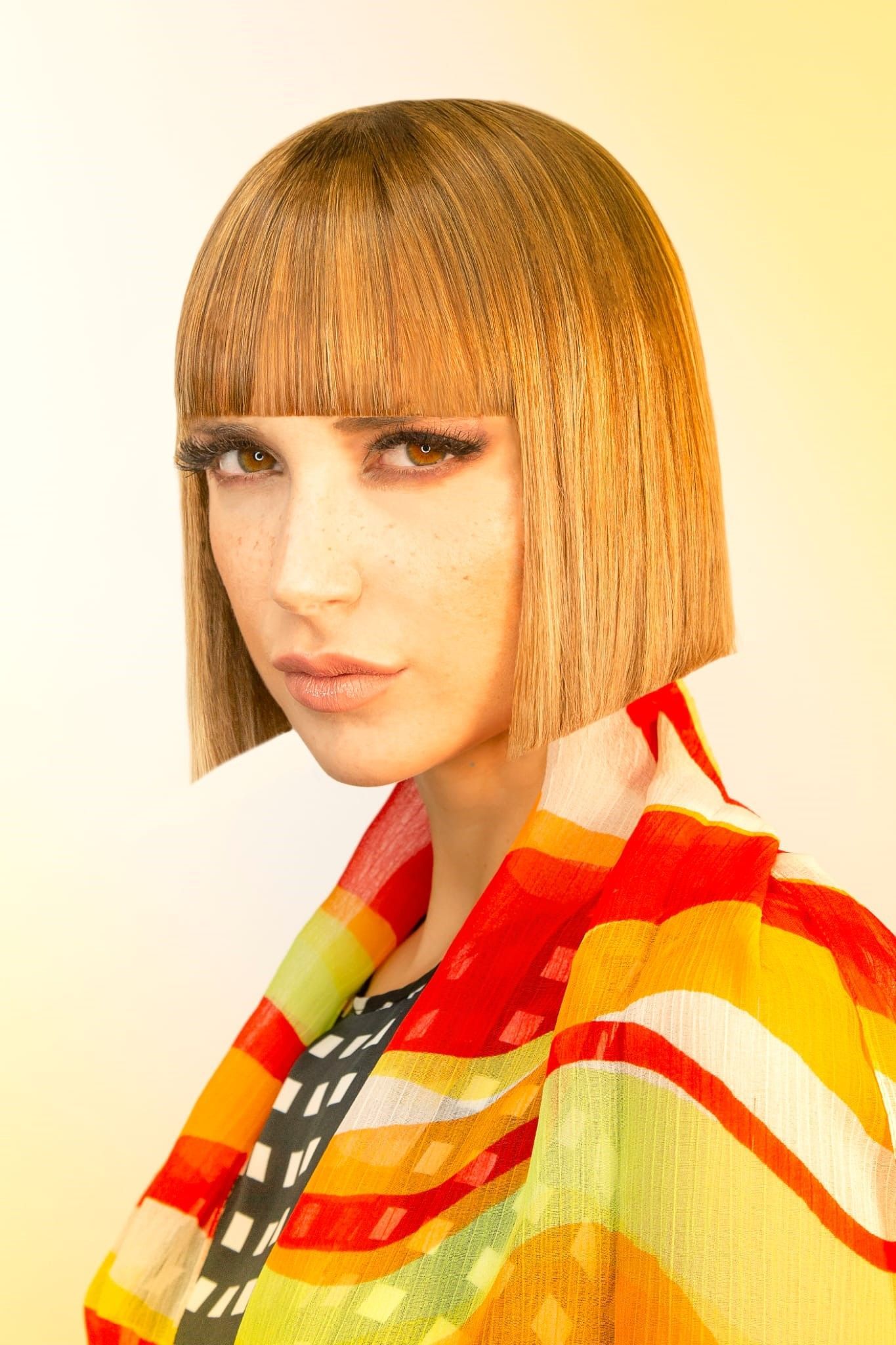 Una persona con capelli biondi, ondulati e tagliati a caschetto, indossa uno scialle a righe vivaci e multicolori su uno sfondo chiaro.