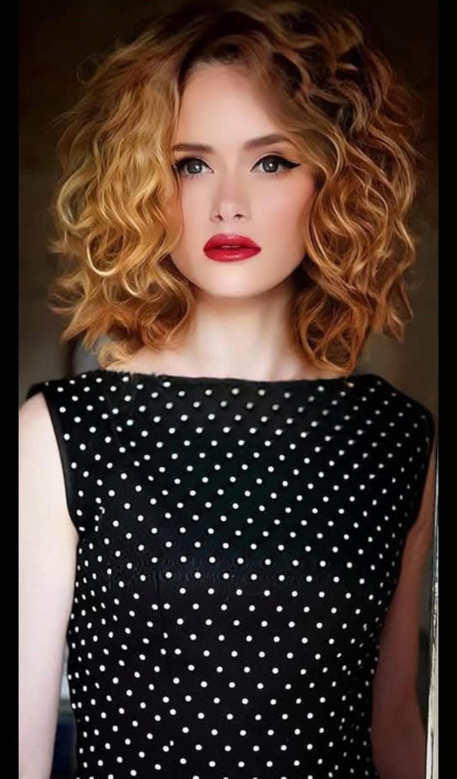Una persona con capelli biondi ricci lunghi fino alle spalle, che indossa una maglietta nera a pois bianchi e un rossetto rosso acceso.