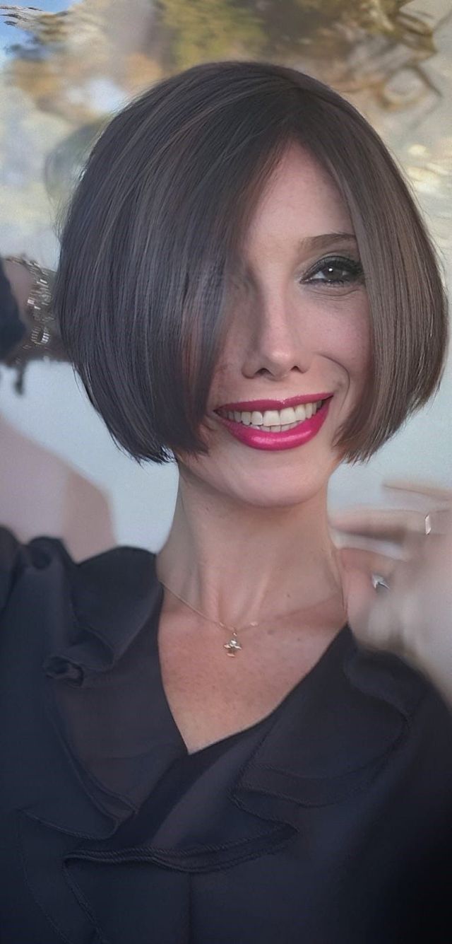 Una persona con un caschetto castano e un rossetto rosa acceso sorride, in posa con una maglietta nera su uno sfondo sfocato.