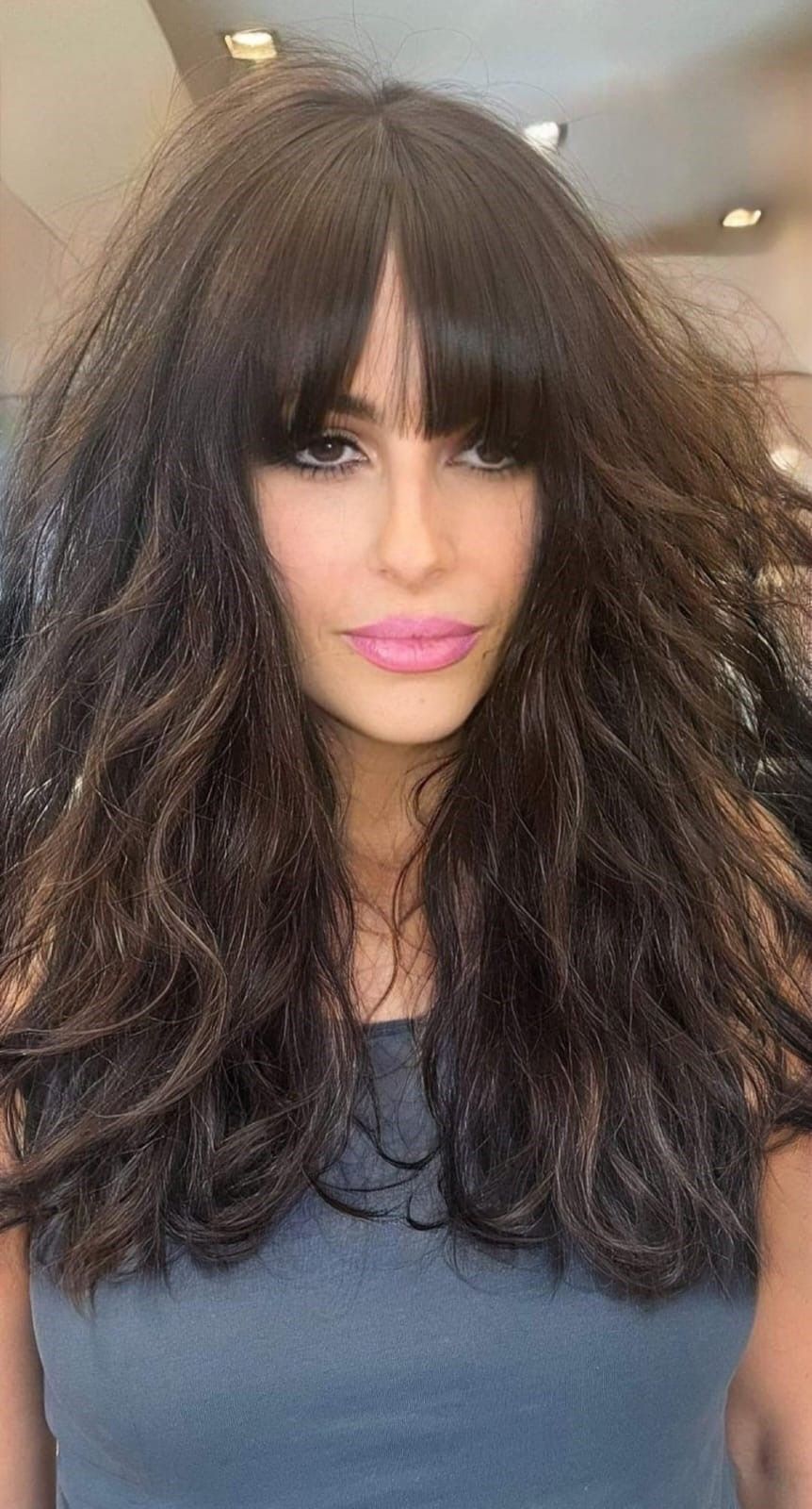 Una persona con lunghi capelli scuri, voluminosi e ondulati, con la frangia, che guarda direttamente in camera con un'espressione neutra.