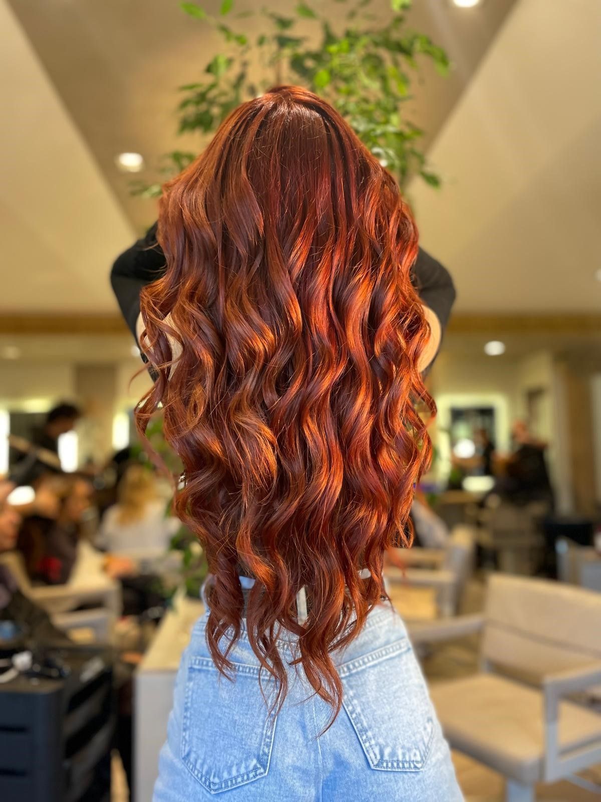 Vista posteriore di lunghi capelli mossi color rame-rosso, acconciati in un salone con sedie e una pianta sullo sfondo sfocato.