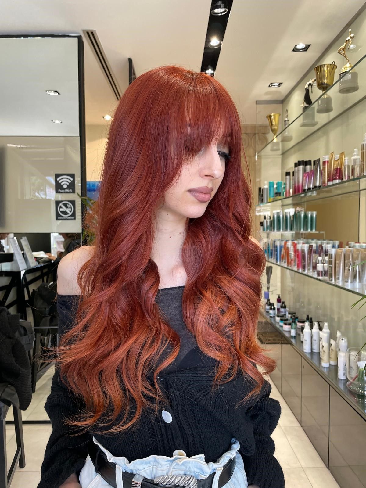 Una persona con lunghi capelli mossi, di un rosso intenso e con la frangia è in piedi in un salone da parrucchiere, con scaffali pieni di prodotti sullo sfondo.