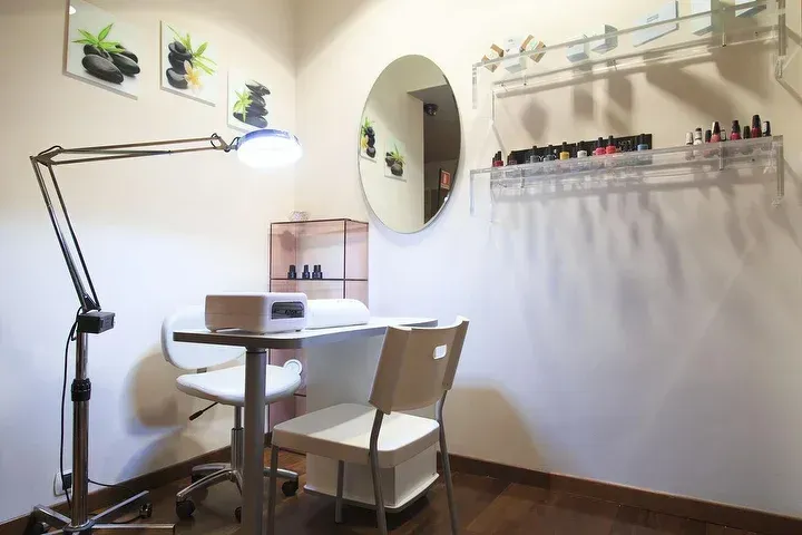 Una piccola postazione minimalista per un salone di bellezza specializzato in unghie, con scrivania bianca, sedia, lampada da lavoro luminosa, specchio e porta smalti a parete.