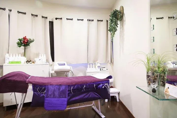 Una stanza da spa con un lettino per trattamenti viola, tende bianche e scaffali che ospitano prodotti per la cura della pelle e piante in vaso.