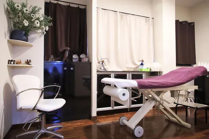 Una sala per trattamenti con una poltrona bianca, un lettino da massaggio con rivestimento viola e mensole addossate a una parete con tenda marrone scuro.