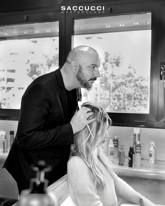 Un parrucchiere con indosso un blazer scuro esamina i capelli biondi di una cliente all'interno di un salone.