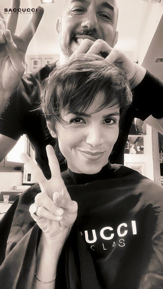 Una persona con i capelli corti sorride e fa il segno della pace, mentre alle sue spalle un parrucchiere fa lo stesso.