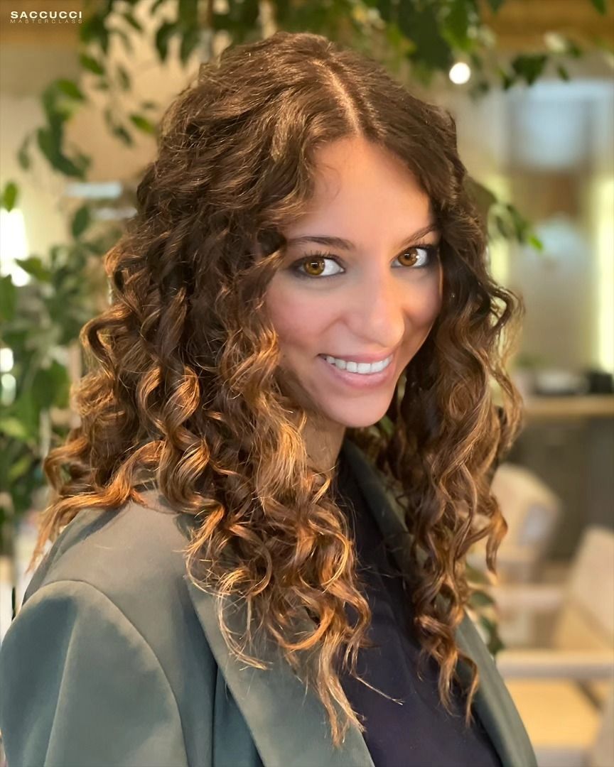 Una persona con lunghi capelli castani ricci e un sorriso smagliante, che indossa un blazer grigio, posa in un ambiente interno illuminato da una luce soffusa.