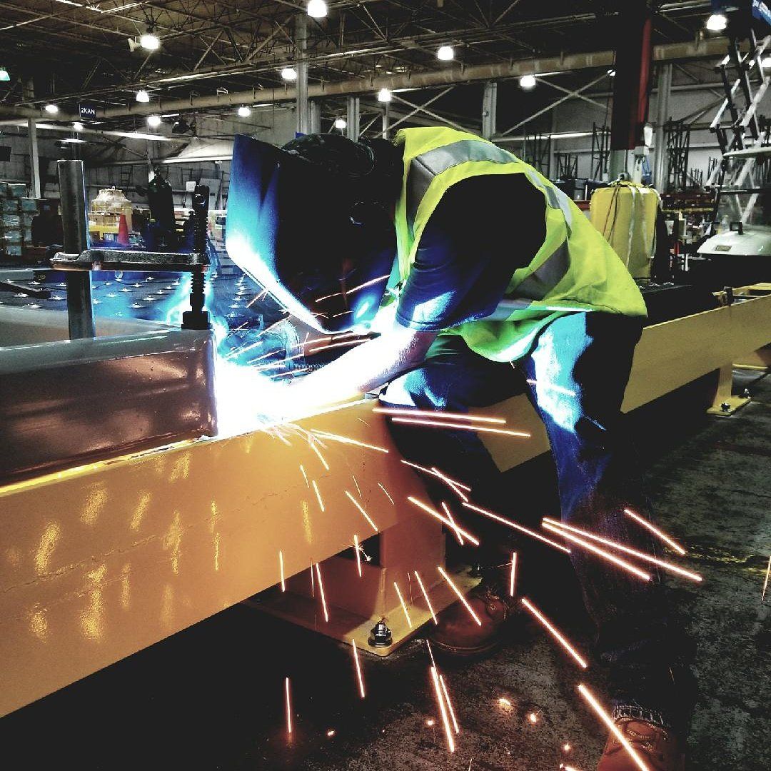 Person welding metal beams in a workshop, sparks flying. They wear a yellow safety vest.