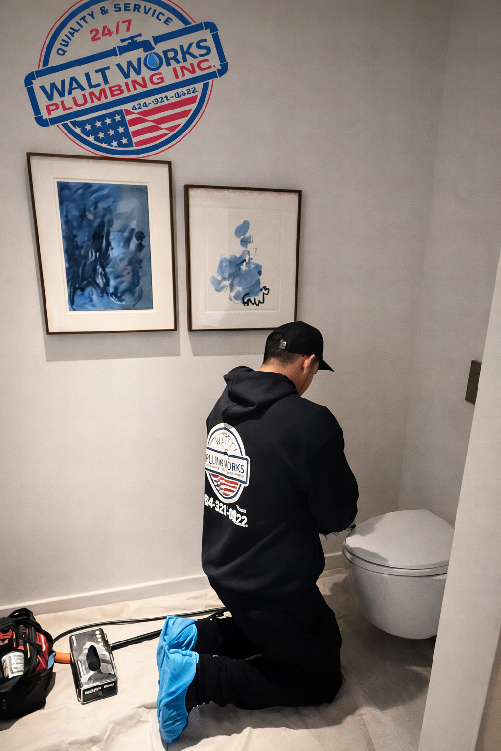 A plumber wearing blue shoe covers kneels to repair a wall-mounted toilet in a room with framed artwork and a company logo.