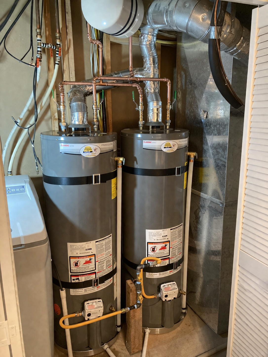 Two grey residential water heaters connected with copper plumbing in a utility closet.