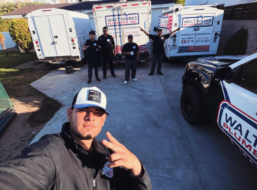 A group of five plumbing company workers stand outside on a concrete driveway near several branded service trucks.