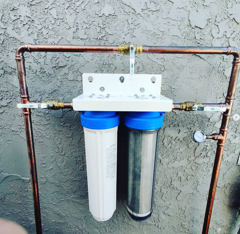 A dual-cartridge water filtration system mounted on a textured wall, connected by copper pipes with multiple shutoff valves.