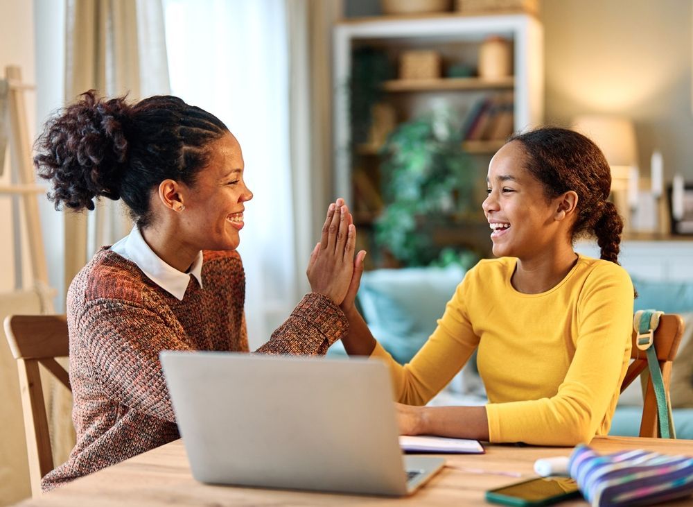A mother and daughter are giving each other a high five while sitting at a table with a laptop.