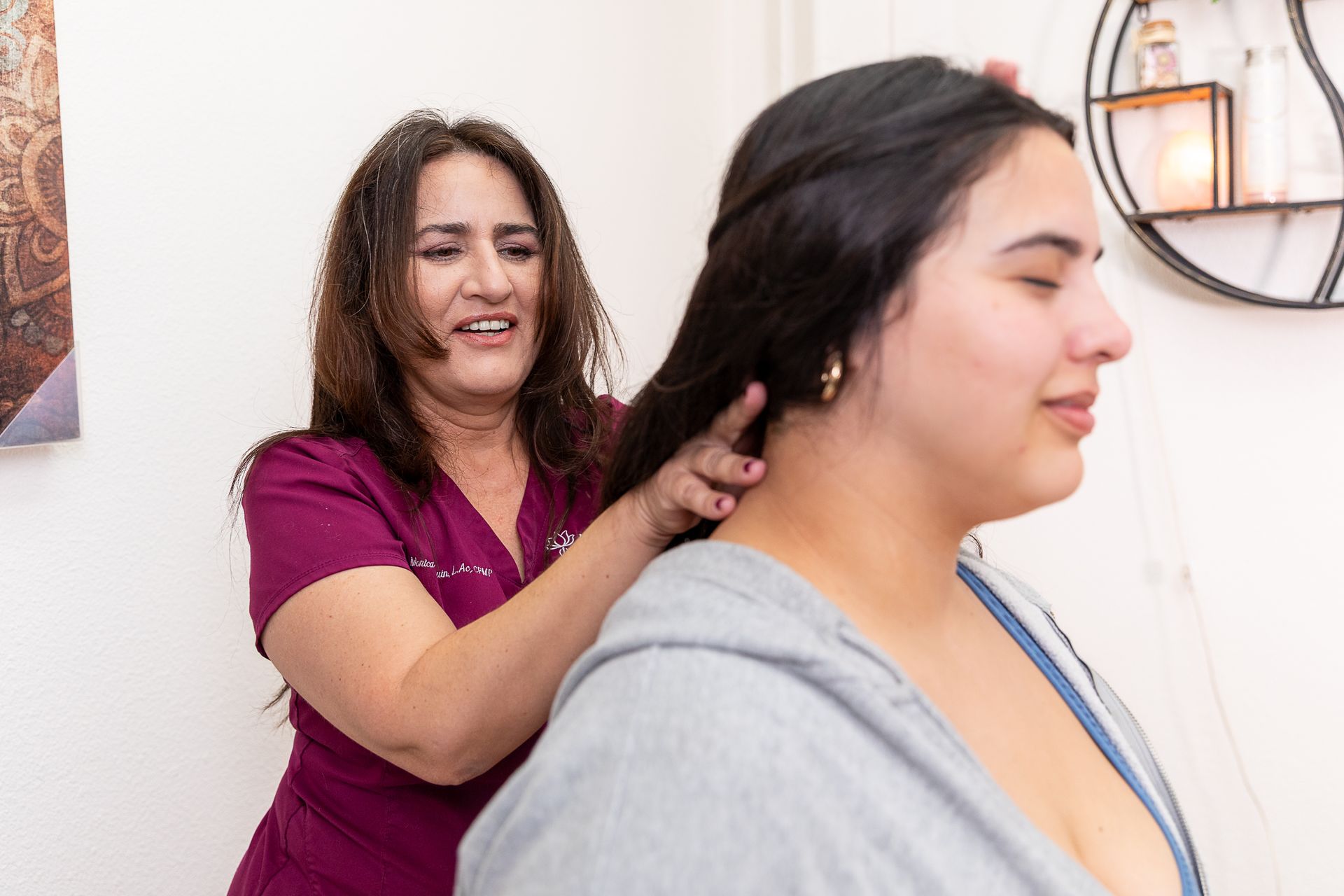 Woman in maroon top massaging the neck of a person in a gray hoodie indoors.