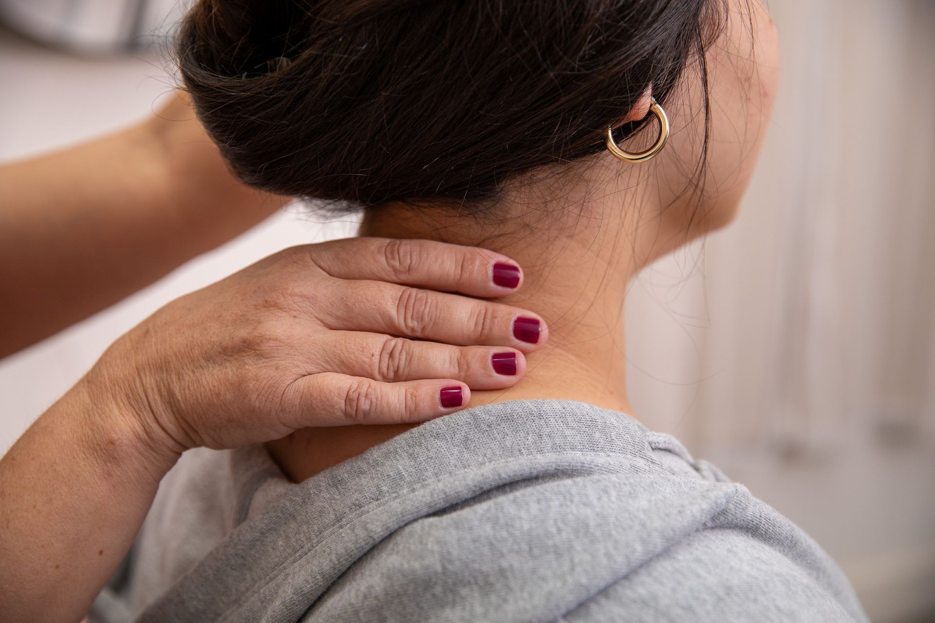 Hands massaging a person's neck, person has dark hair up, wearing a grey sweater and gold earring.