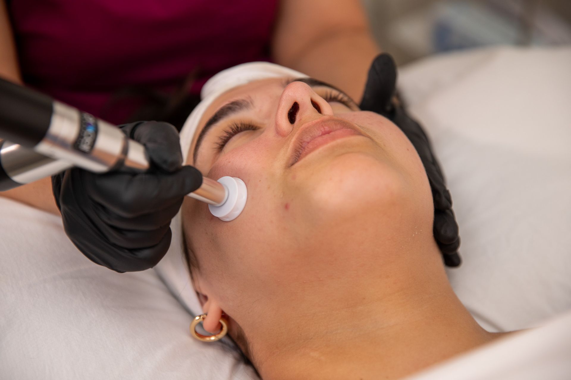 Woman receiving facial treatment with device, indoors. Dark gloves, white towel, focused expression.